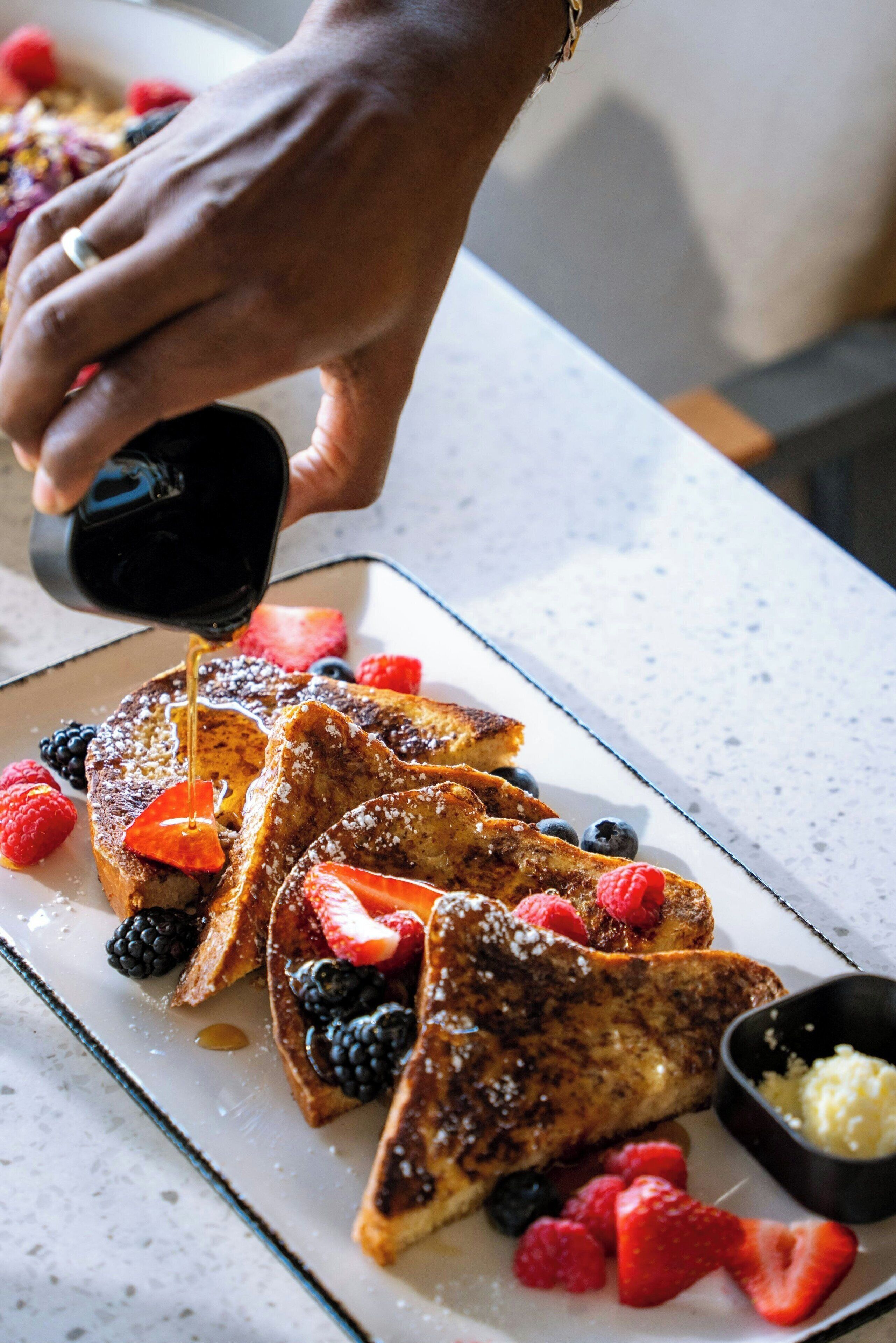 A hand pouring syrup over French toast with berries and a small bowl of butter.
