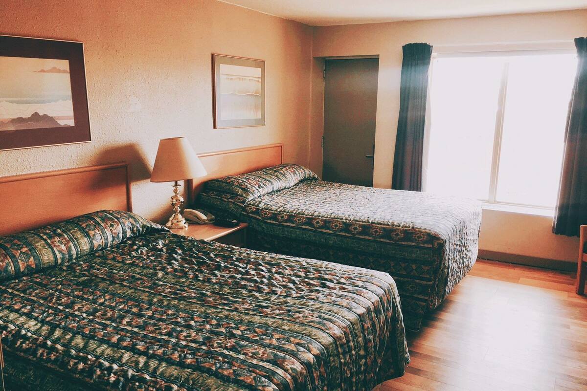 A hotel room with two beds, a nightstand, a lamp, and a window with curtains.