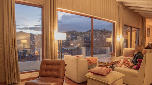 Amplia sala de estar con un ventanal que ofrece vista a la ciudad, un sillón de cuero, un sofá y una mesita con lámpara.