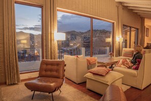 A spacious living room with a large window offering a city view, a leather chair, a sofa, and a small table with a lamp.