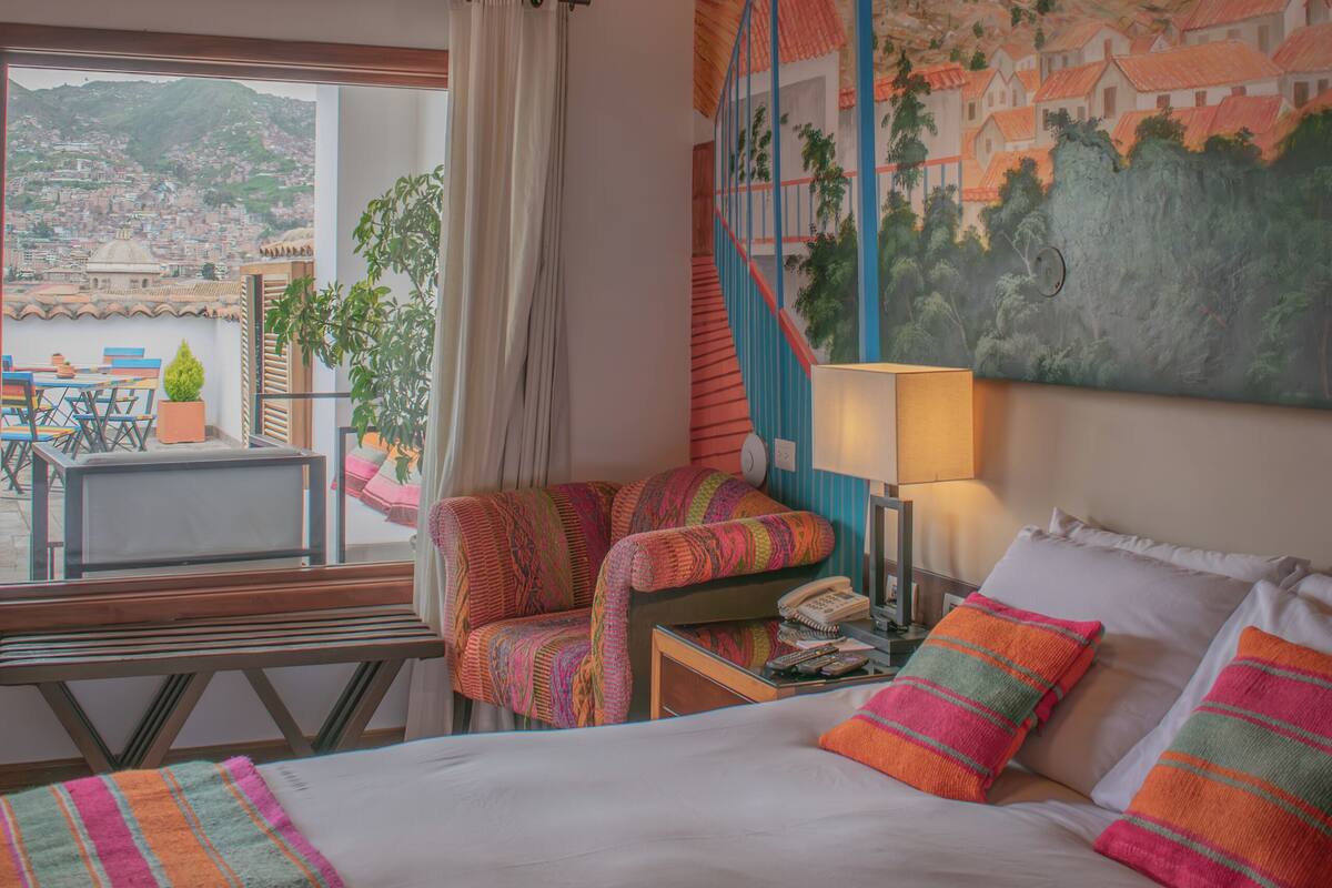 Une chambre à coucher avec un lit, des oreillers, une lampe, une chaise, une table et une vue sur le paysage urbain par la fenêtre.