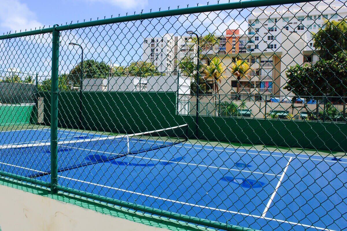 A tennis court with a chain-link fence, surrounded by buildings and greenery.