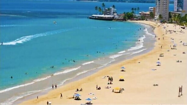 Une plage avec une eau d’un bleu limpide, un rivage sablonneux et quelques bâtiments en arrière-plan.