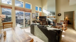 A spacious living room with a large sofa, a wooden dining table, and a fireplace. There are multiple windows providing a view of the mountains.