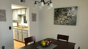 A dining area with a dark wooden table, four chairs, and a fruit bowl. There is a kitchen visible in the background. - Tomar Park Apt 2 - KP101APT2 (Sioux Falls)