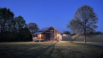 A rustic barn with a porch and a covered area, surrounded by trees and open fields.