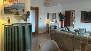 Ein Wohnzimmer mit einer grünen Konsole, einem beigen Sofa, einem Essbereich mit Tisch und Stühlen sowie einem Gemälde einer Küstenlandschaft.
