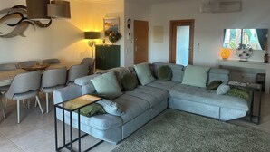 Ein modernes Wohnzimmer mit einem grauen Ecksofa, einem Esstisch mit Stühlen, einer Stehlampe und einem Fernseher auf einem weißen Schrank.