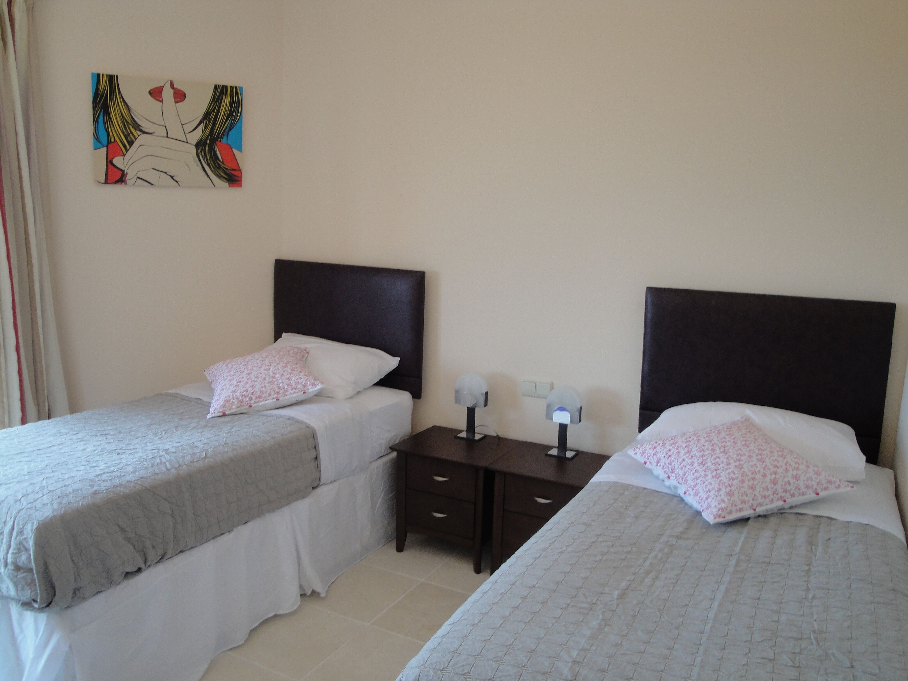 A hotel room with two single beds, each with a headboard, a bedside table with a lamp, and a wall-mounted artwork.