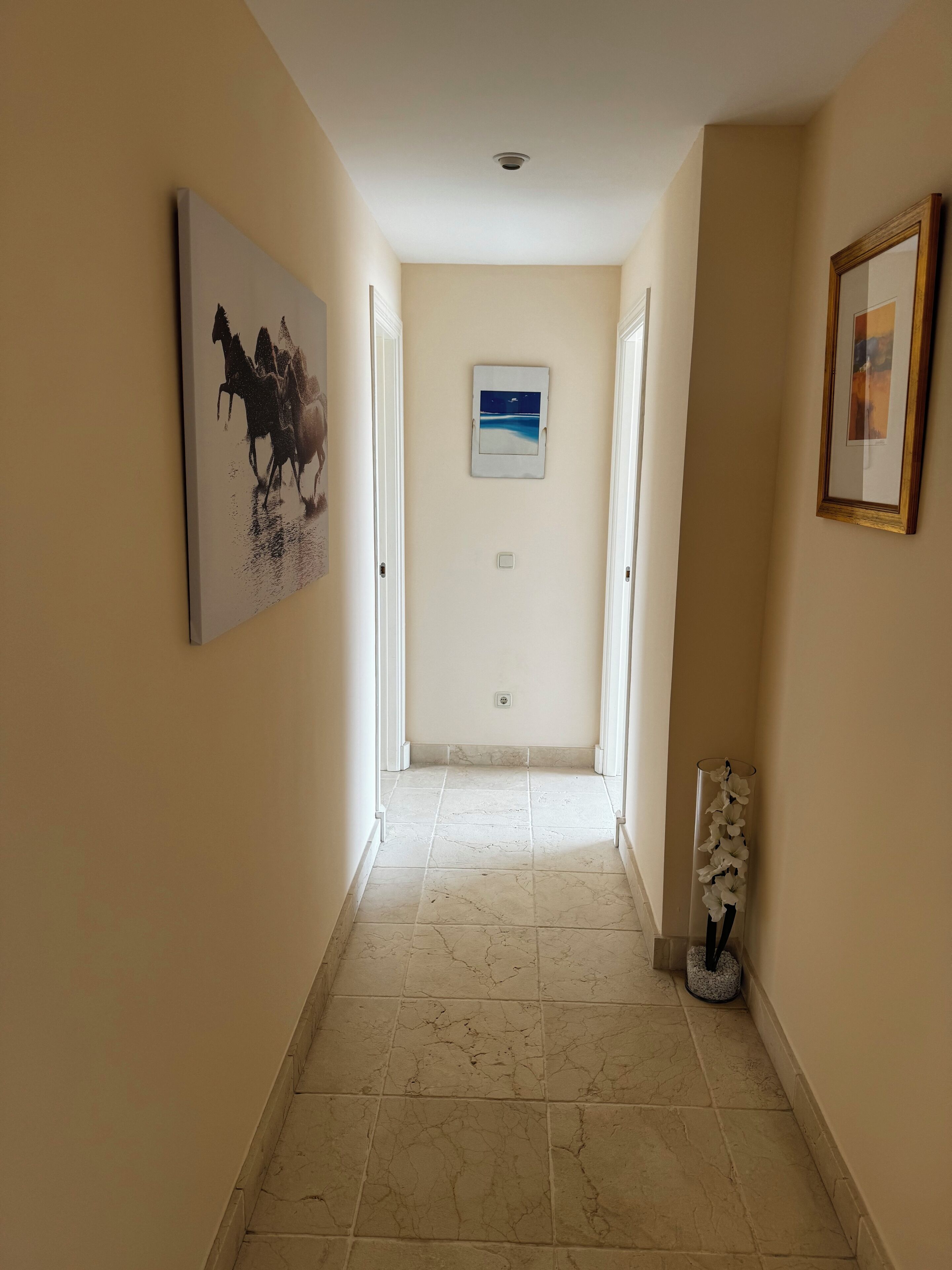 A hallway with a tiled floor, a framed picture on the left, a picture on the wall, and a picture frame on the right.