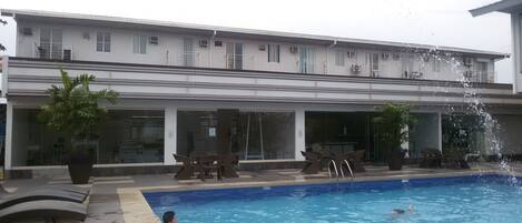 Una piscina con una fuente de agua, sillas de descanso y un edificio con múltiples ventanas y balcones al fondo.