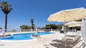 Un área de piscina con sillones de descanso y sombrillas, rodeada por una cerca blanca y palmeras.