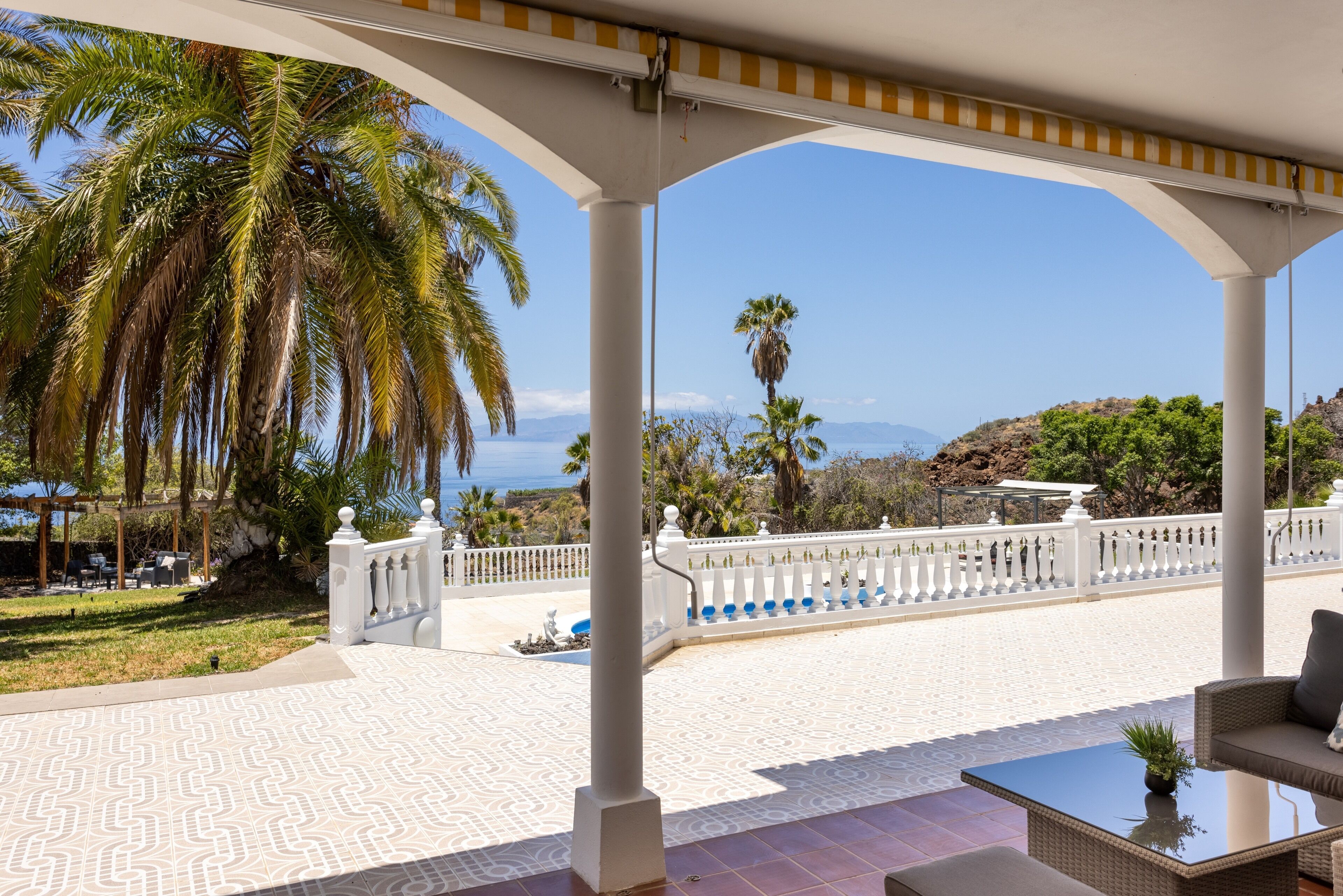 Un espacio al aire libre cubierto con piso estampado, columnas blancas y vistas al mar y colinas.