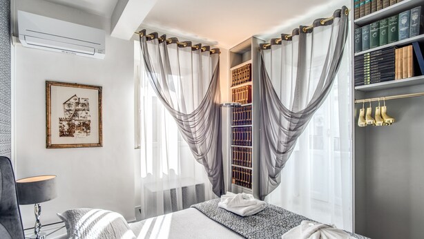 Una habitación de hotel con una cama, una mesita de noche con lámpara, una estantería con libros y una ventana con cortinas.