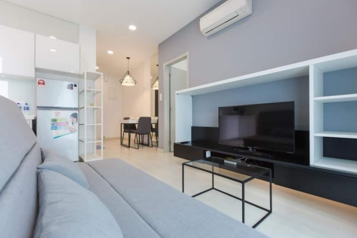 A modern living room with a grey sofa, a flat-screen TV mounted on a black wall unit, and a dining area with a table and chairs.