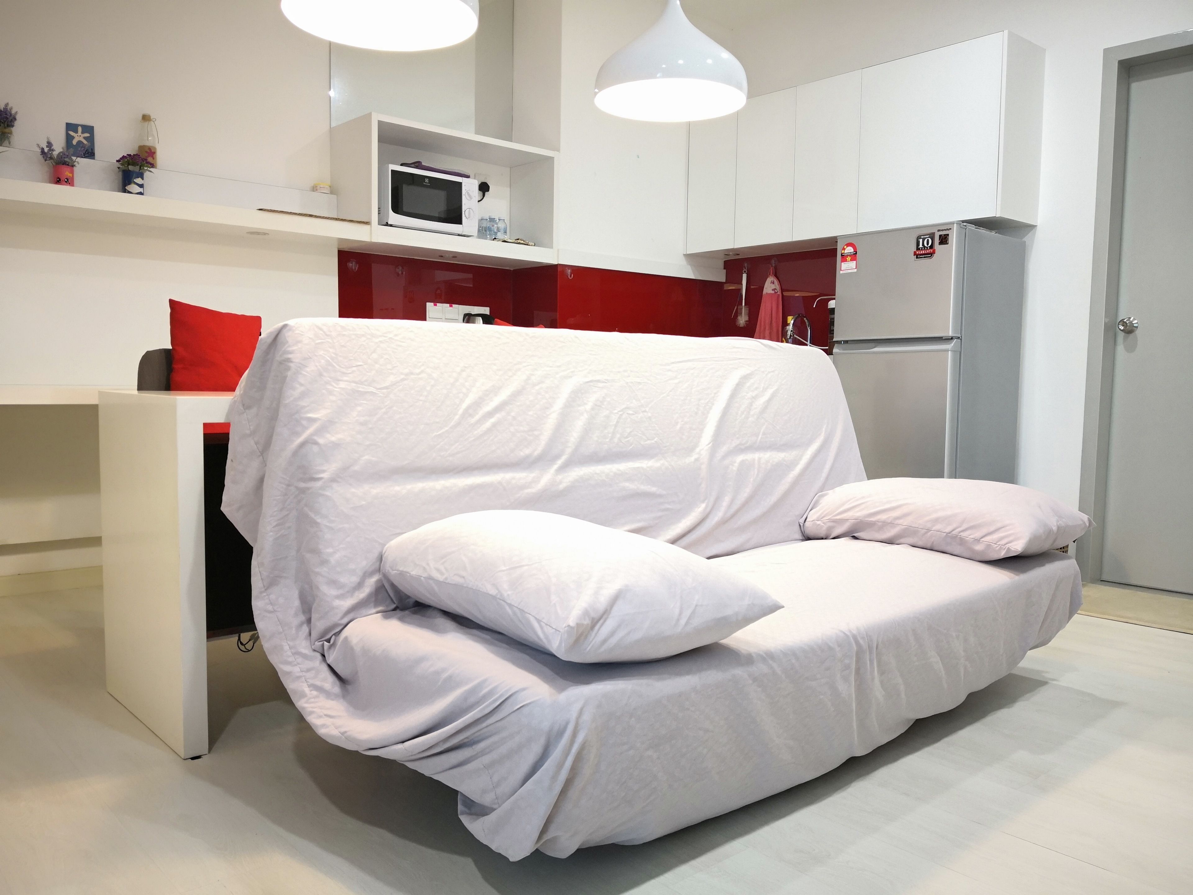 Un salon moderne avec un canapé blanc, un mur d’accent rouge et un coin cuisine doté de meubles blancs et d’appareils électroménagers blancs.
