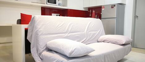 Una sala de estar moderna con un sofá blanco, una pared de acento roja y una zona de cocina con armarios y electrodomésticos blancos.
