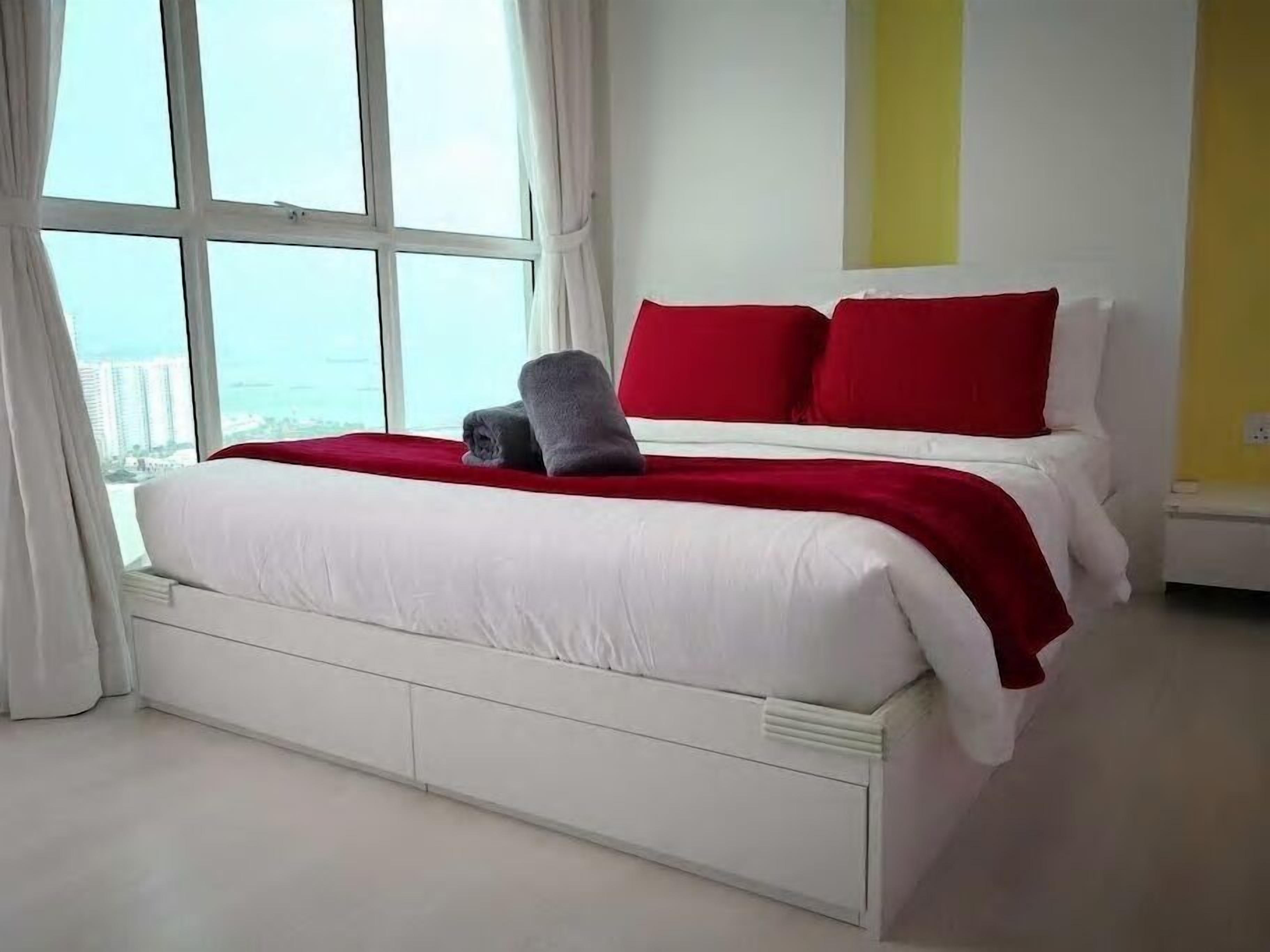Une chambre d’hôtel avec une grande fenêtre, un lit avec une literie blanche et des oreillers rouges, et une vue sur la ville.