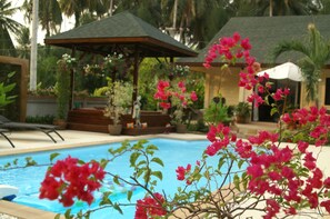Un área de piscina con un pabellón, rodeada de exuberante follaje y flores rosadas.