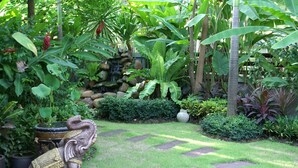 Een weelderige tuin met een stenen olifantbeeld, een kleine waterval en diverse tropische planten.