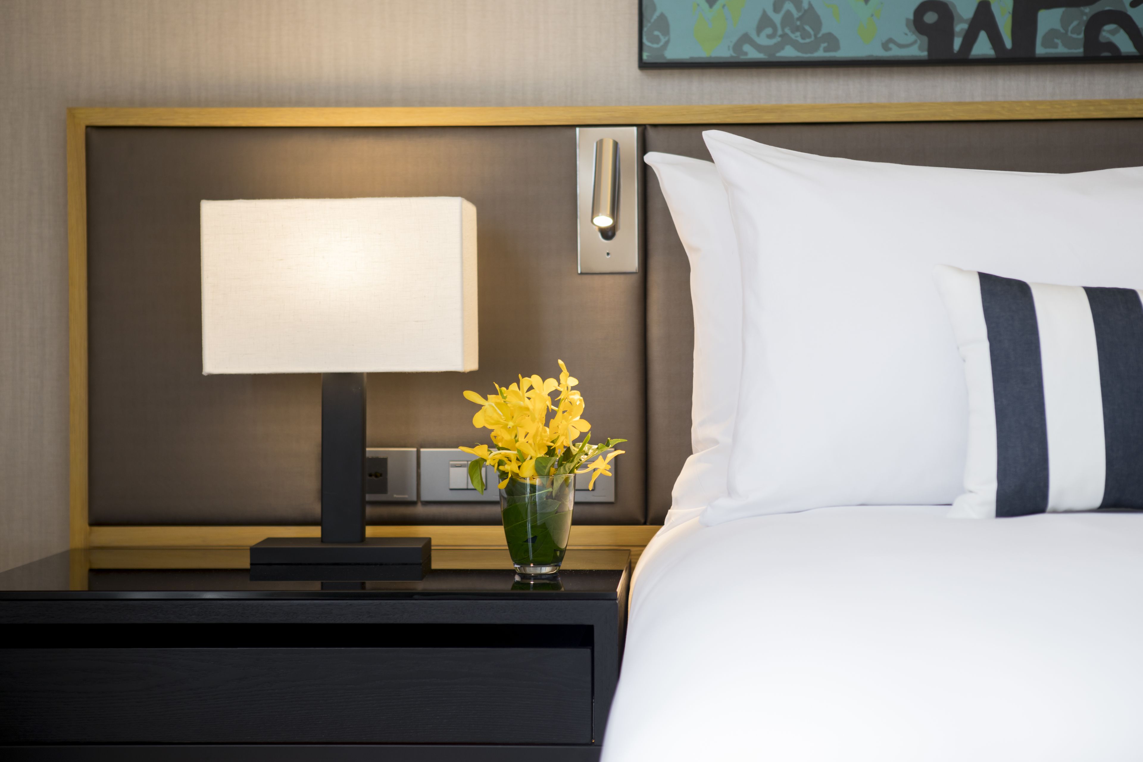 A neatly made hotel room with a bed, a bedside table with a lamp, and a vase of flowers.