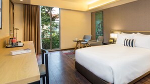 A hotel room with a large bed, a desk with a chair, a small table with two chairs, and a view of greenery through a window.