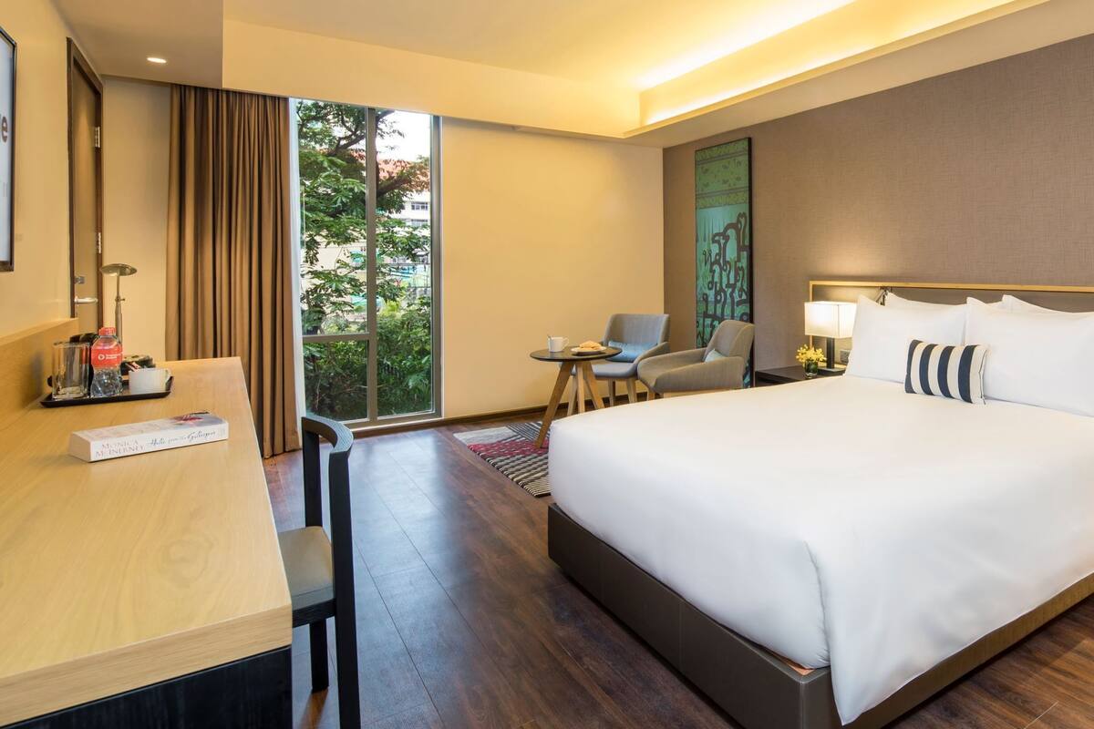 A hotel room with a large bed, a desk with a chair, a small table with two chairs, and a view of greenery through a window.
