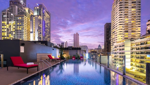 A rooftop infinity pool with red lounge chairs, surrounded by high-rise buildings and a city skyline at dusk.