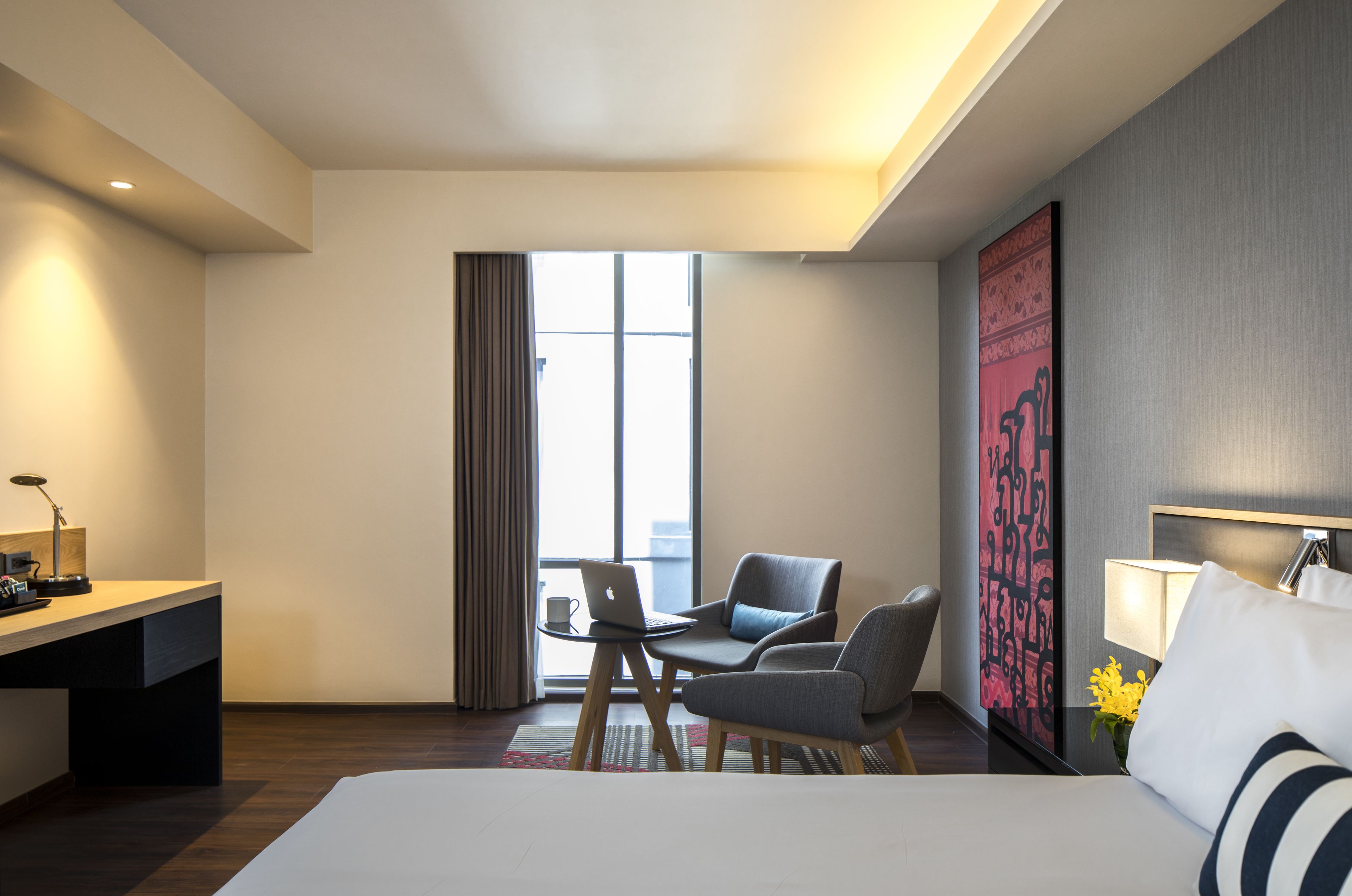 A hotel room with a bed, a desk, two chairs, a laptop, a lamp, and a large window with curtains.