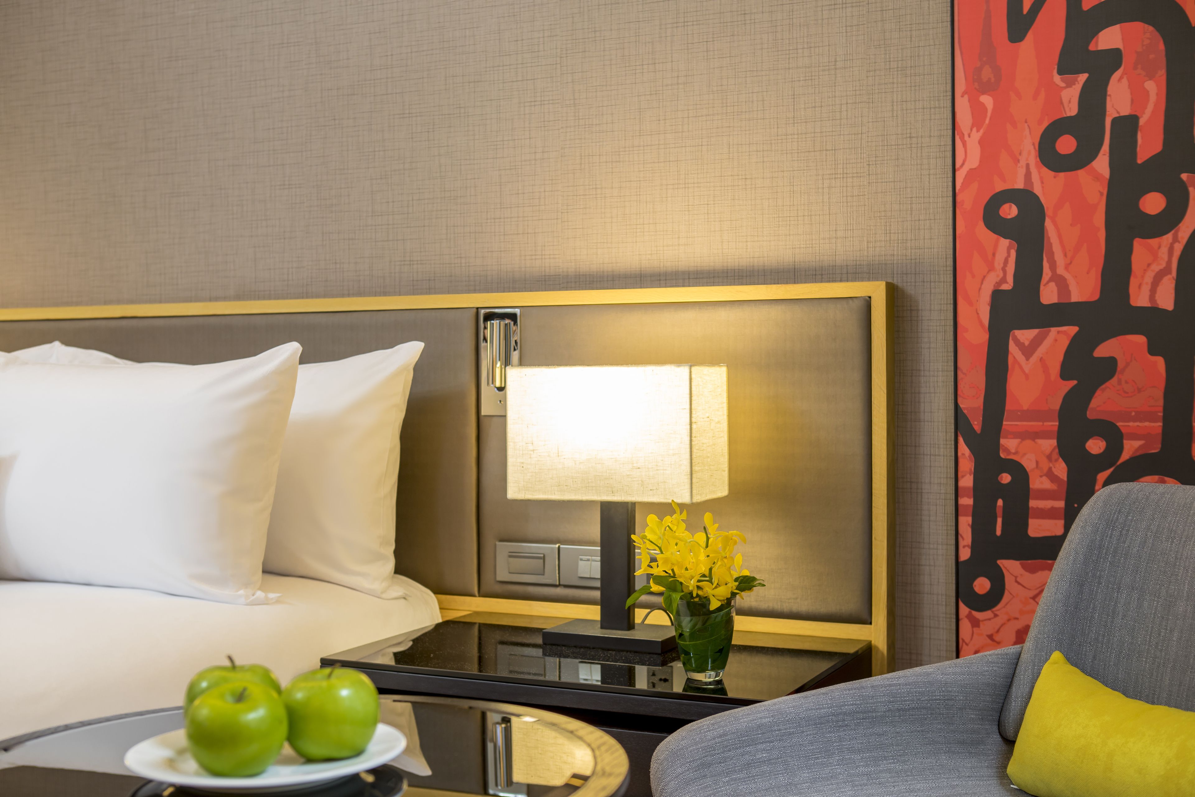 Habitación de hotel con cama, mesita de noche con lámpara, un jarrón con flores y un plato con manzanas.