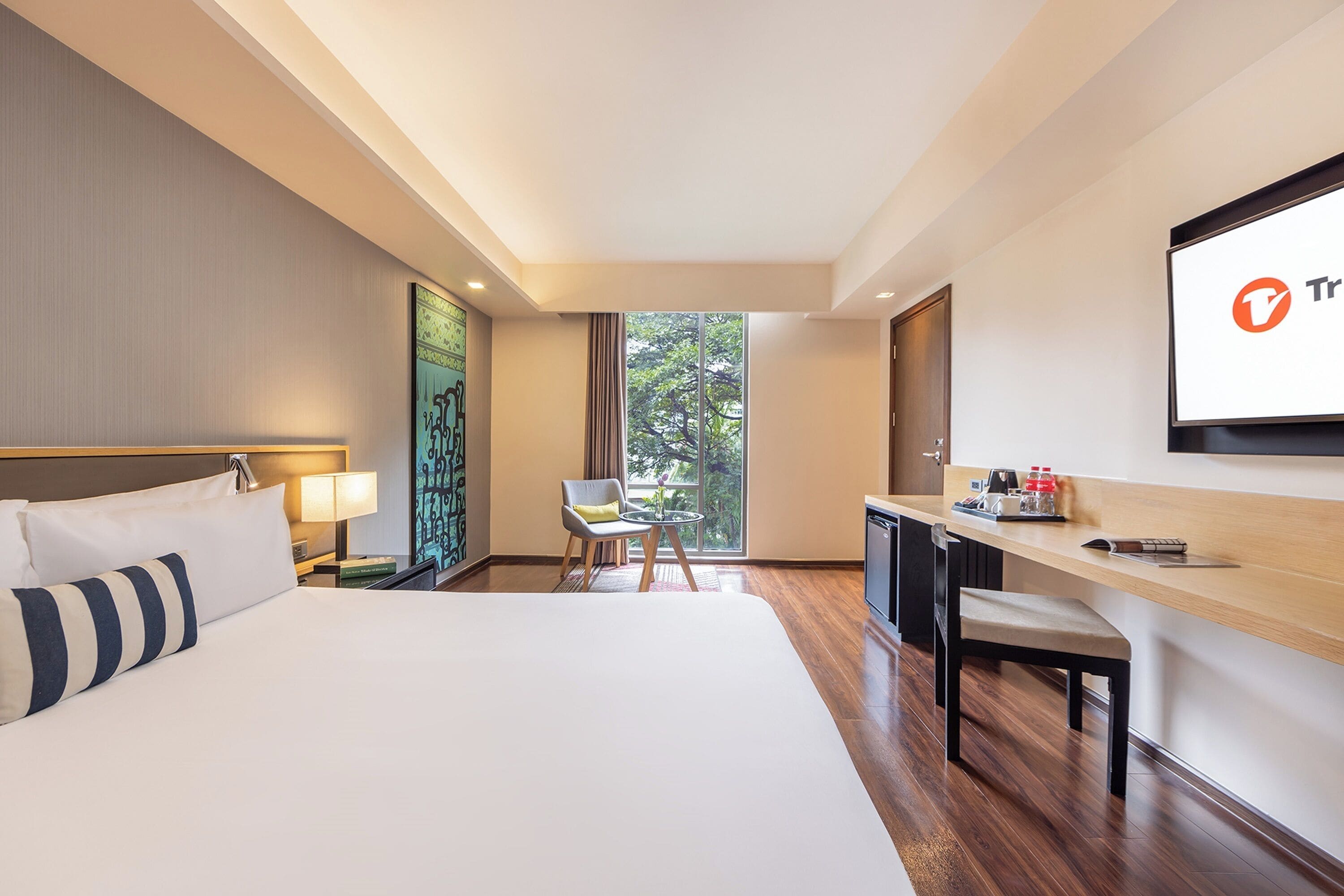A hotel room with a large bed, a desk, a chair, a TV, and a window with a view of trees.