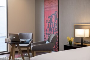 Ein Hotelzimmer mit einem Bett, einem Schreibtisch mit einem Laptop, einem Sessel, einem Nachttisch mit einer Lampe und einer Wandverkleidung.
