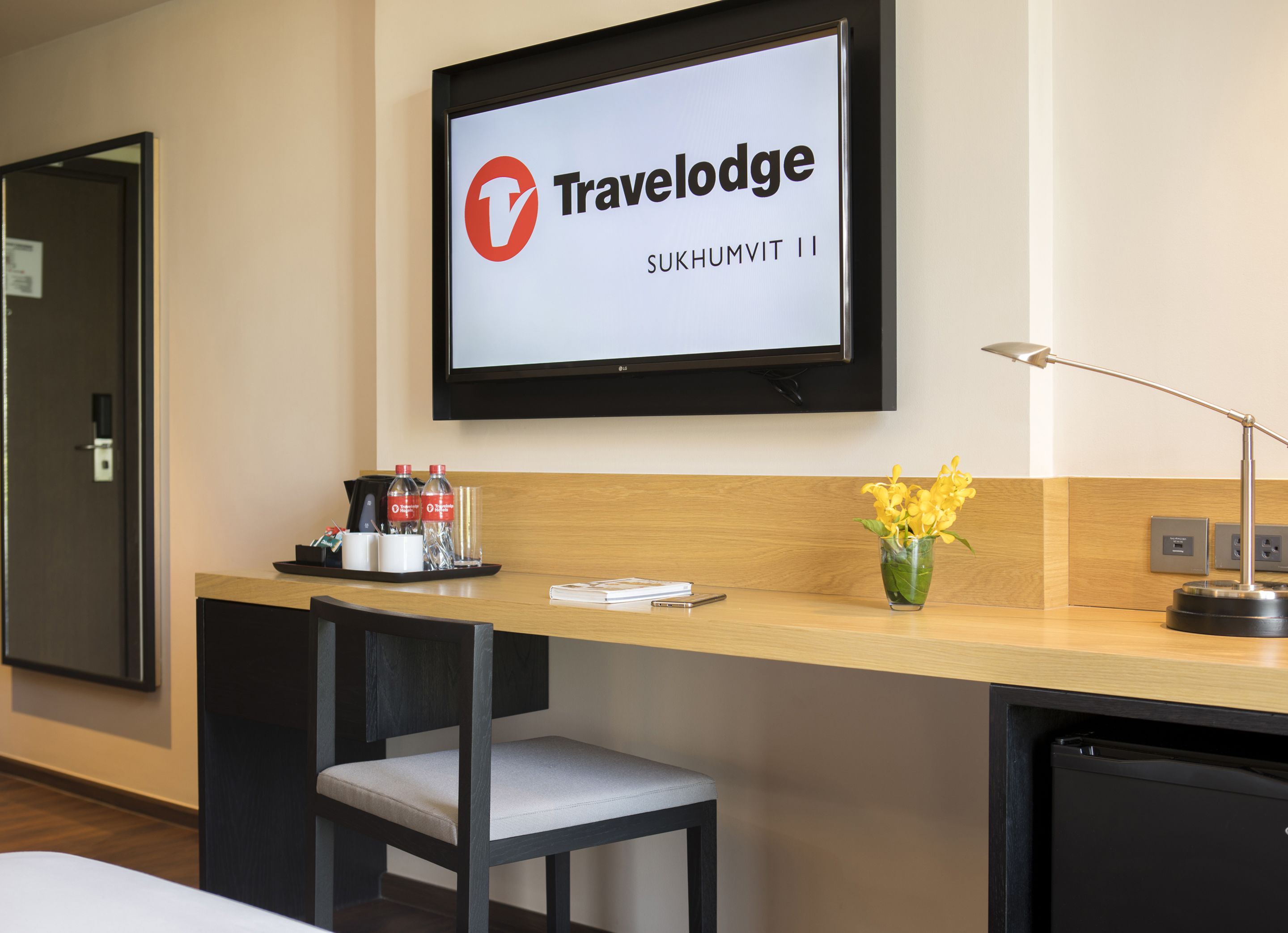 A hotel room with a bed, a desk with a computer monitor, a chair, a small table with a vase of flowers, and a wall-mounted TV displaying the Travelodge logo.