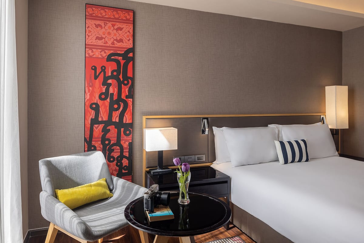 A hotel room with a bed, a chair, a nightstand, a lamp, and a decorative wall piece.