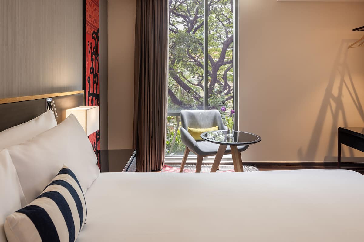A hotel room with a large bed, a chair, a small table, and a view of trees through a window.
