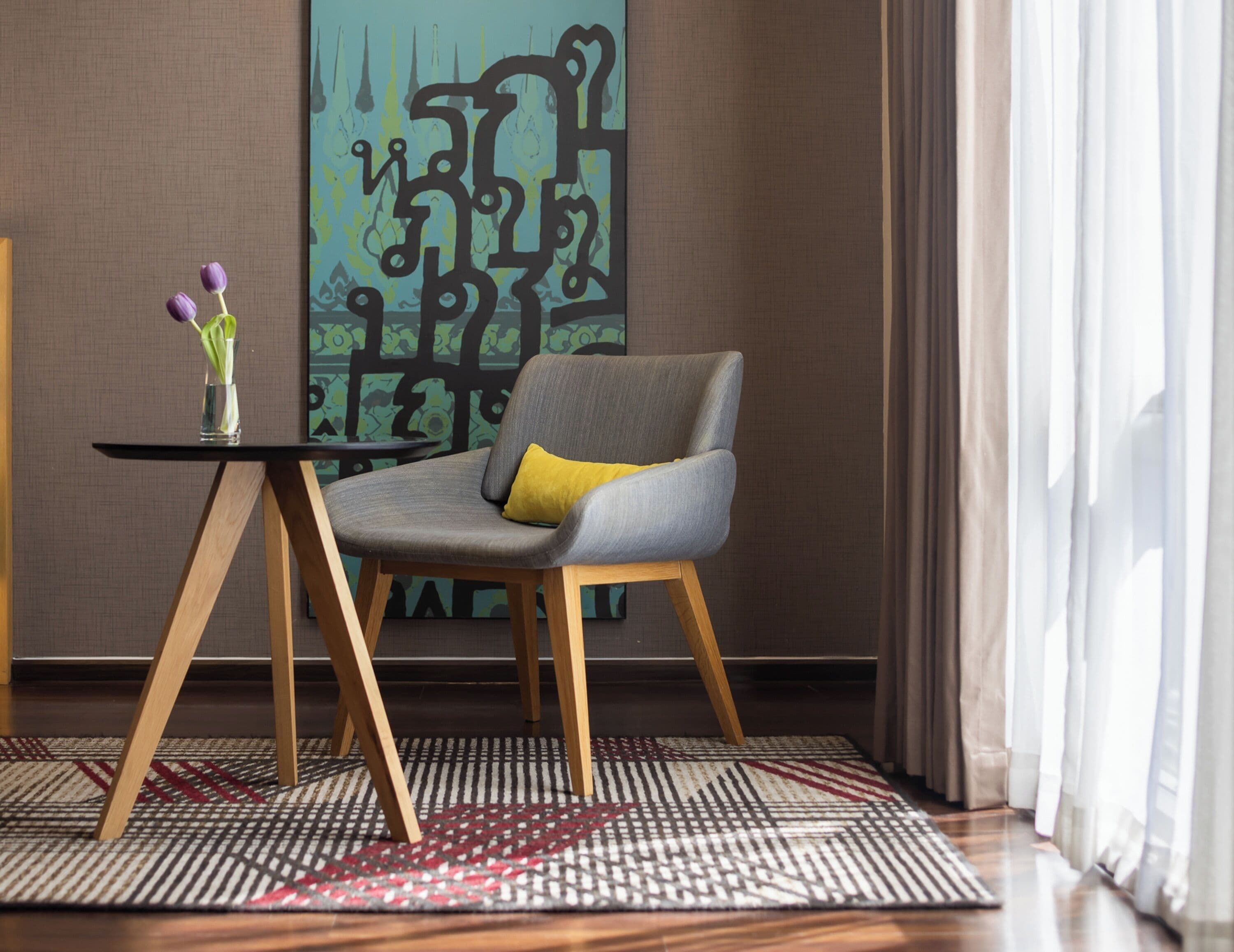 A modern room with a grey armchair, a small table with a vase of flowers, and a patterned rug.