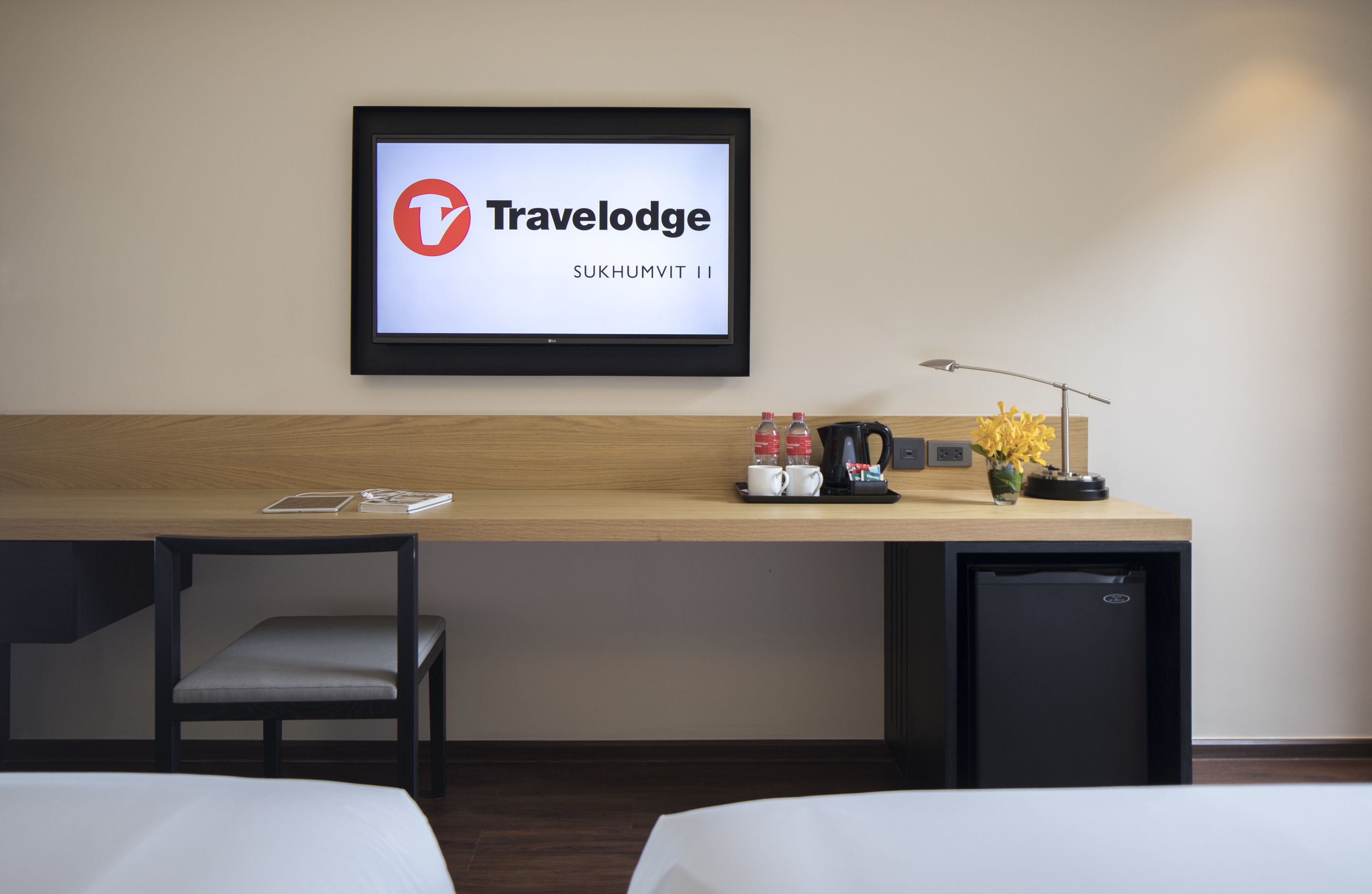 Hotelkamer met een bed, een bureau met een laptop, een stoel, een kleine koelkast en een televisie waarop het Travelodge logo staat.