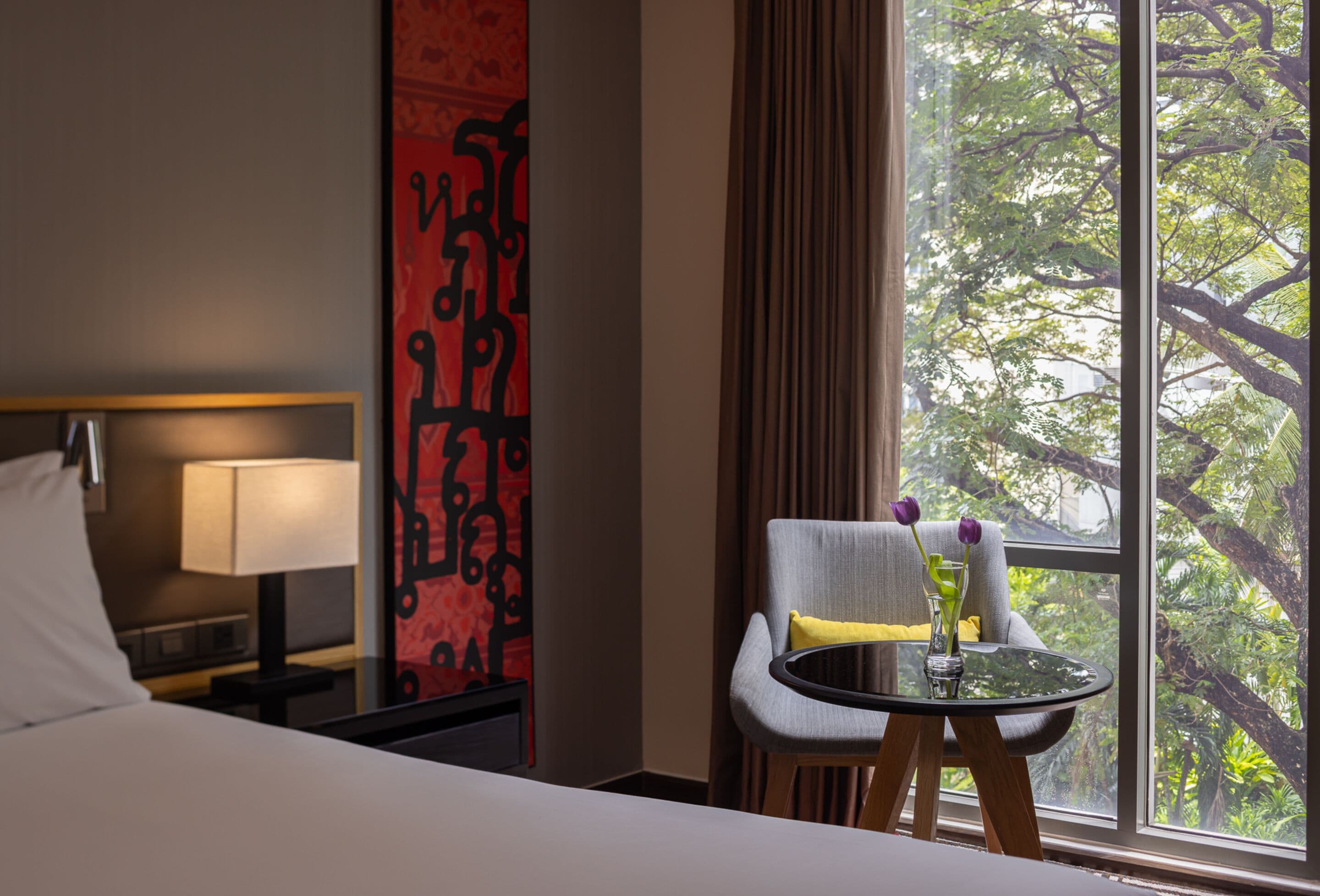 Une chambre d’hôtel avec un lit, une table de chevet avec une lampe, une chaise agrémentée d’un vase de fleurs et une fenêtre donnant sur des arbres.