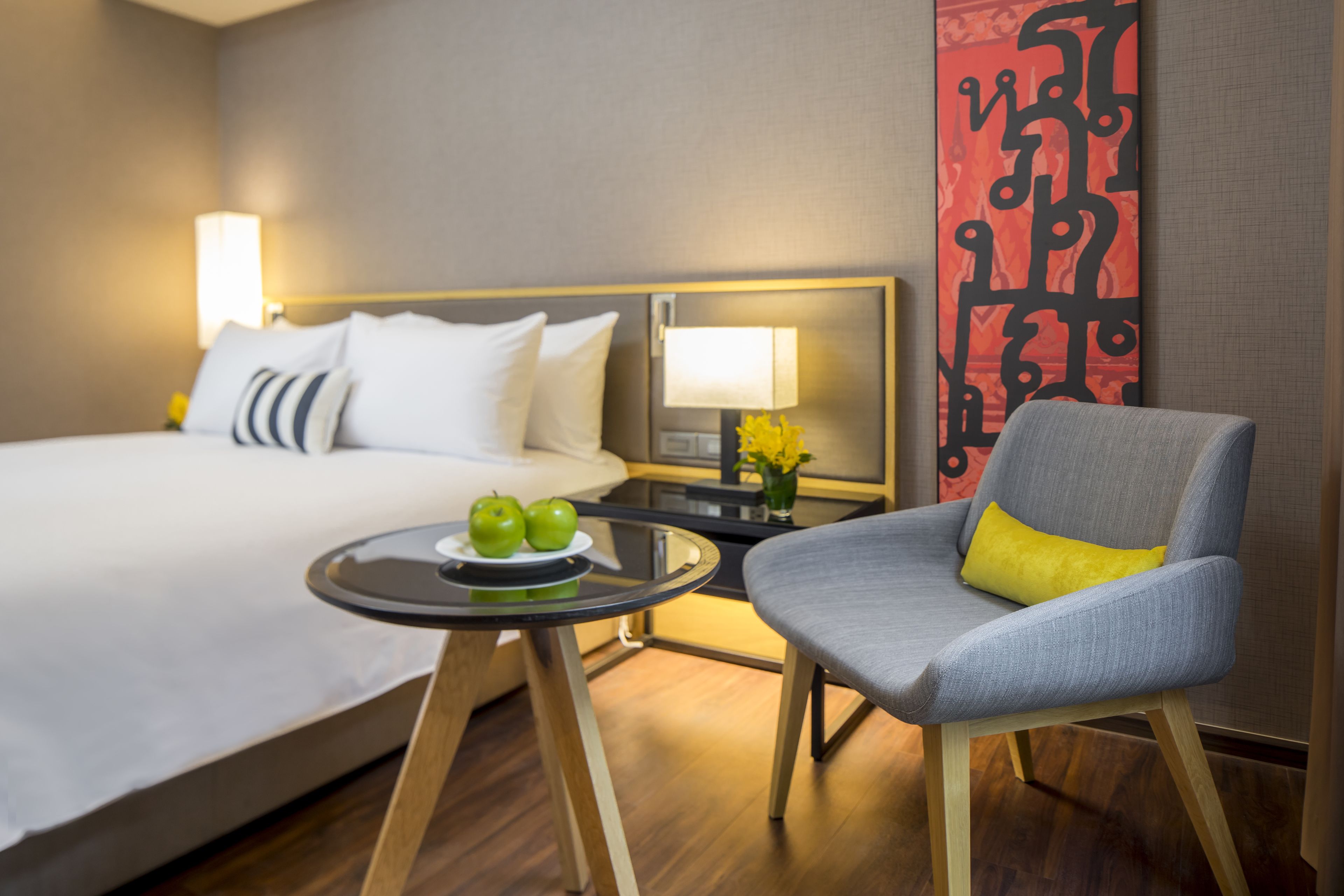 A hotel room with a bed, a small table with apples, a chair, and a lamp.