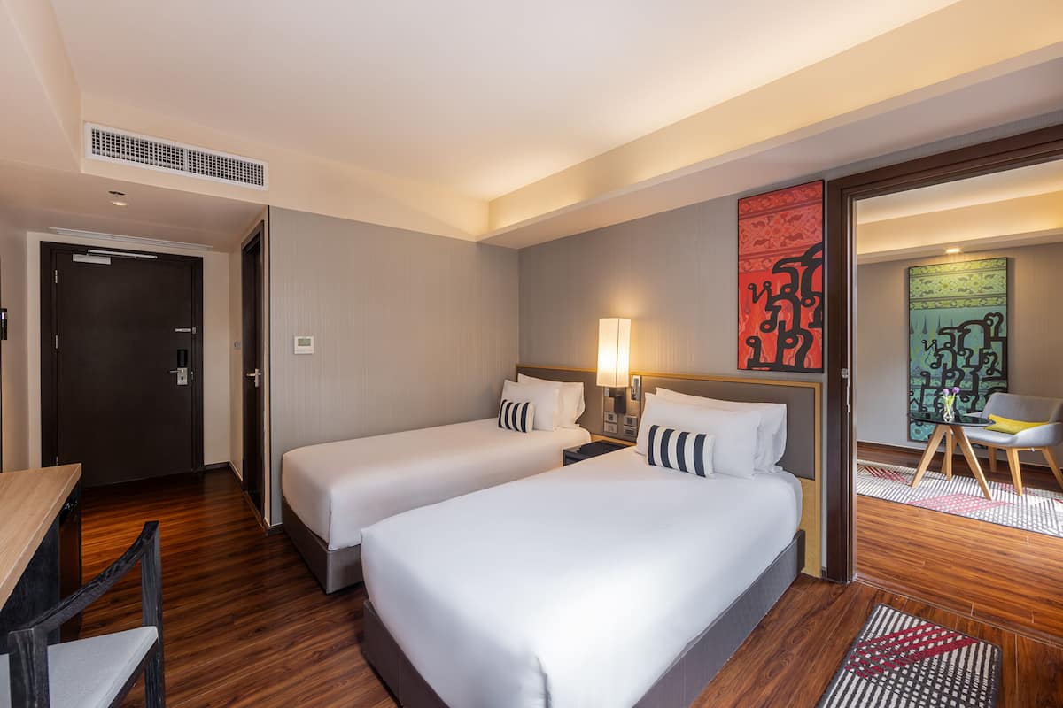 A hotel room with two beds, a desk, a chair, a lamp, and artwork on the wall.
