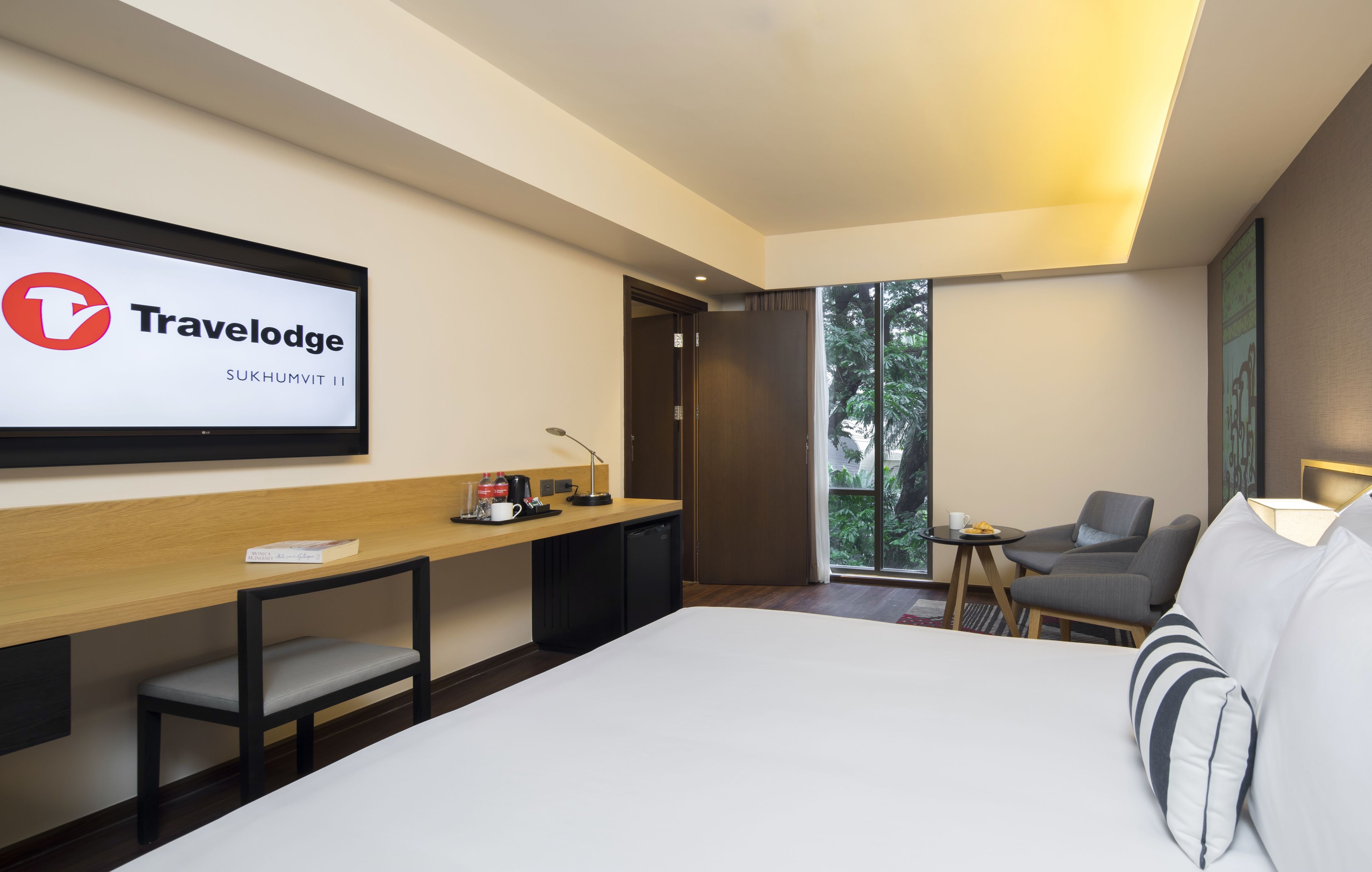 Ett modernt hotellrum med en stor säng, ett skrivbord med en stol, ett litet bord och en soffa. På väggen sitter en tv som visar Travelodges logotyp.