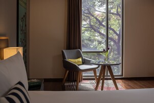 Hotelkamer met een groot raam, een stoel met een geel kussen, een klein rond tafeltje met een vaas, en een bed met een gestreept kussen.