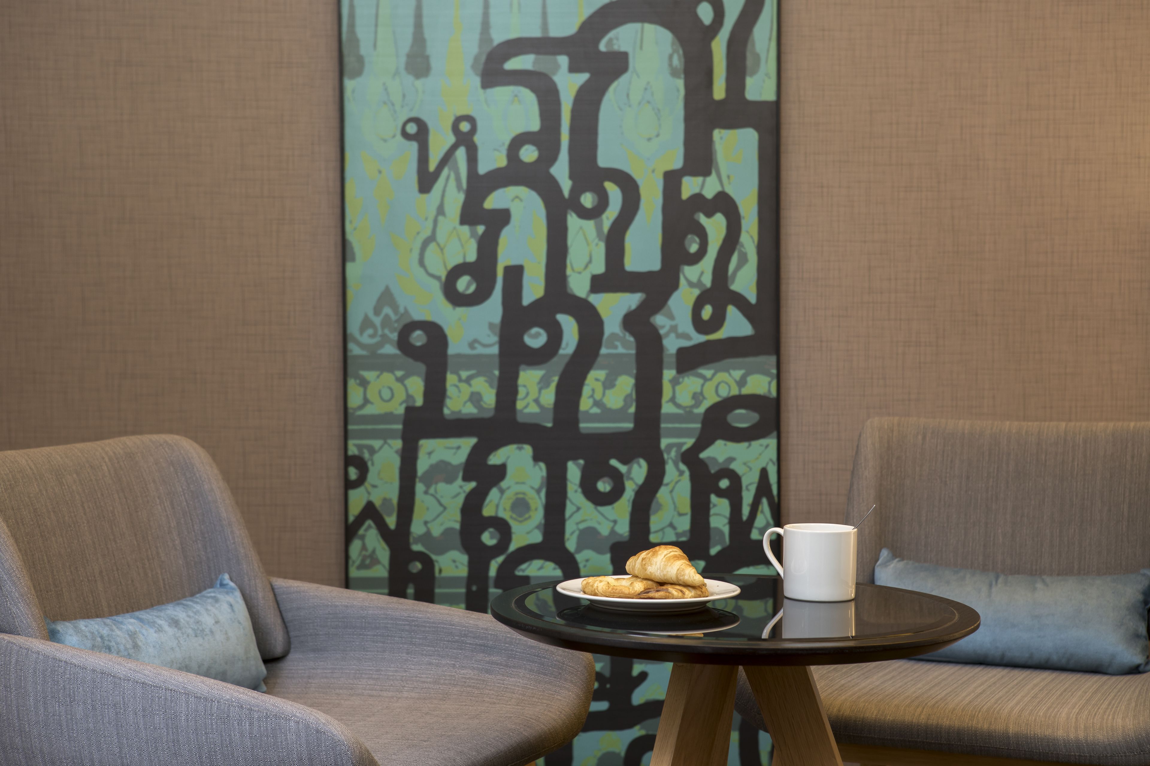 A room with two armchairs, a small table with a plate of croissants and a cup, and a decorative wall with abstract patterns.