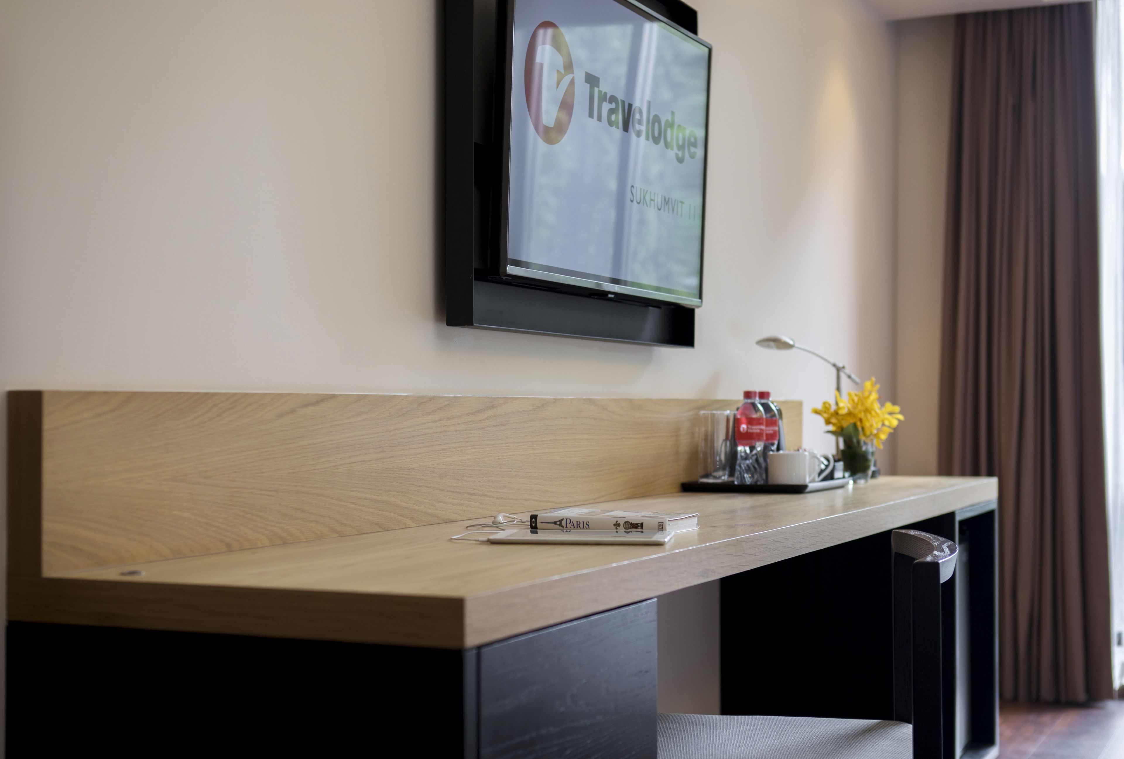 A modern reception desk with a mounted TV displaying the Travelodge logo, a wooden counter, and a small table with a vase of yellow flowers.