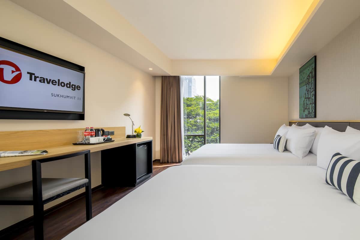 A modern hotel room with a large bed, a desk with a computer, a TV displaying the Travelodge logo, and a window with a view of trees.