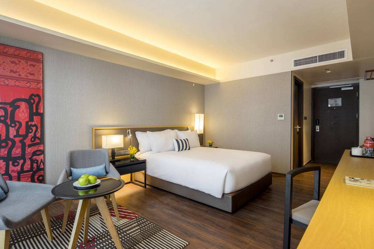 A hotel room with a bed, a chair, a small table with apples, a lamp, and a red abstract painting on the wall.