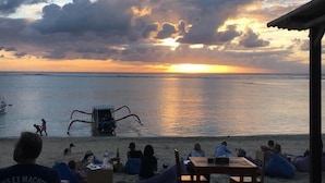 A beach scene with people sitting on bean bags and a table, a boat in the water, and a sunset in the background. - Linda Beach Hotel (Lembongan Island)