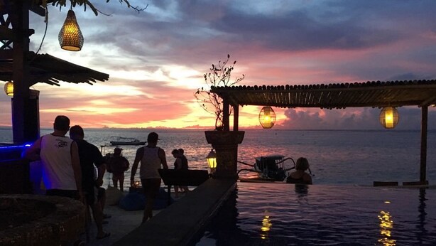 A sunset view from a poolside area with a covered seating area and a boat in the background. - Linda Beach Hotel (Lembongan Island)