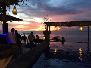 A sunset view from a poolside area with a covered seating area and a boat in the background. - Linda Beach Hotel (Lembongan Island)