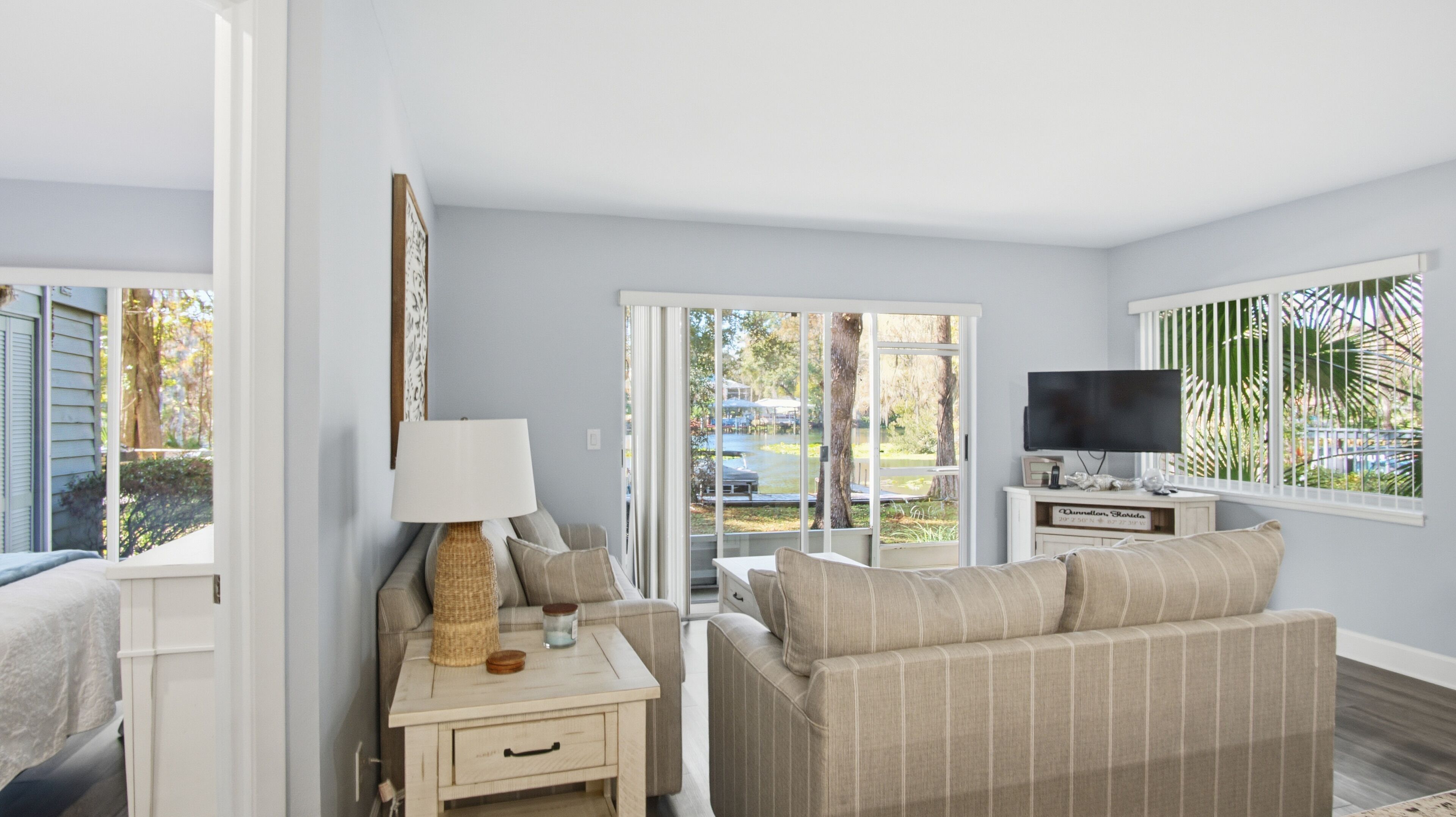 A room with a sofa, a TV, a lamp, and a window with a view of a pool and trees.